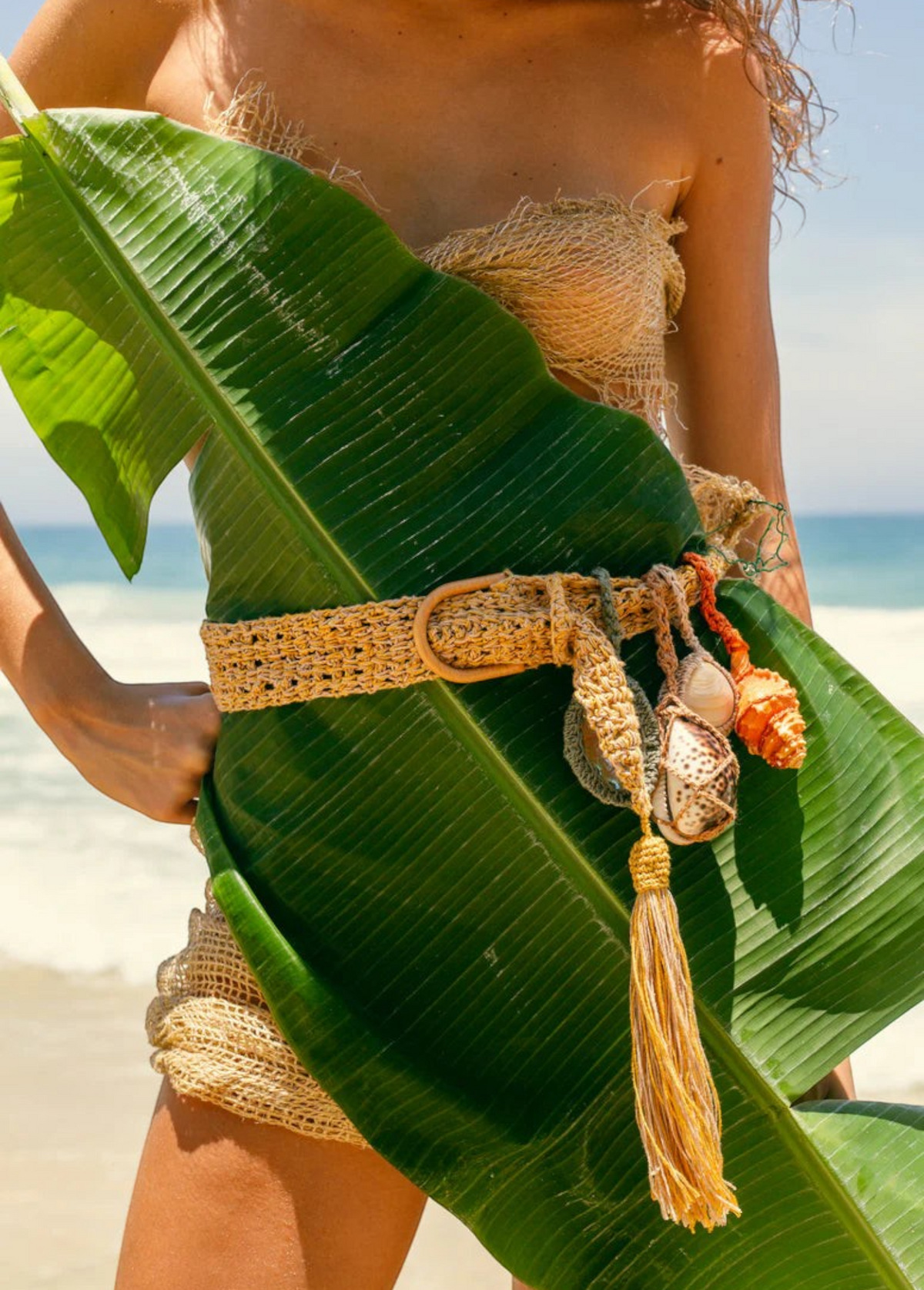 KABE SHOP INC Jibe belt in natural woven fiber with removable seashell charms and tassel, worn on a beach.