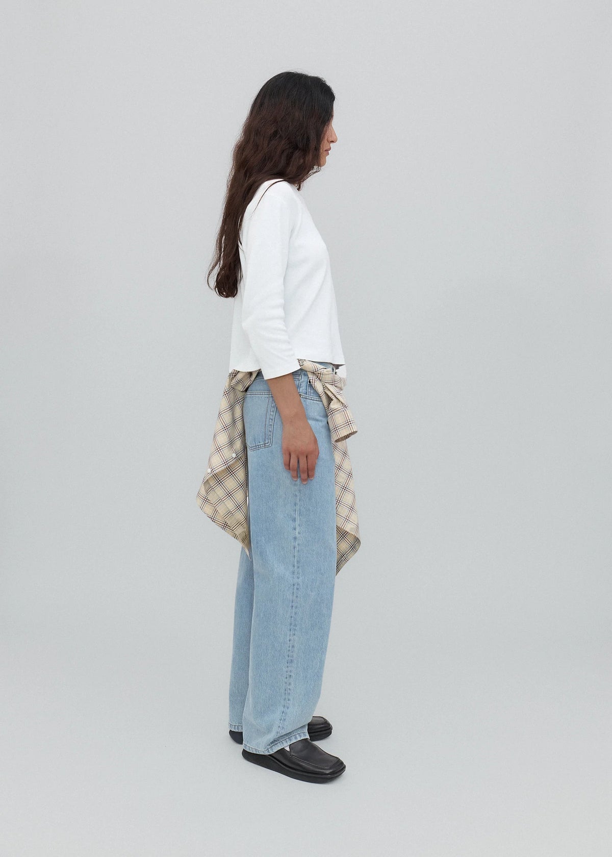 A person stands sideways wearing a white long-sleeve top, black shoes, and light blue Everyday Jeans with a relaxed silhouette. A plaid shirt is tied around their waist, and the background is plain and light gray.