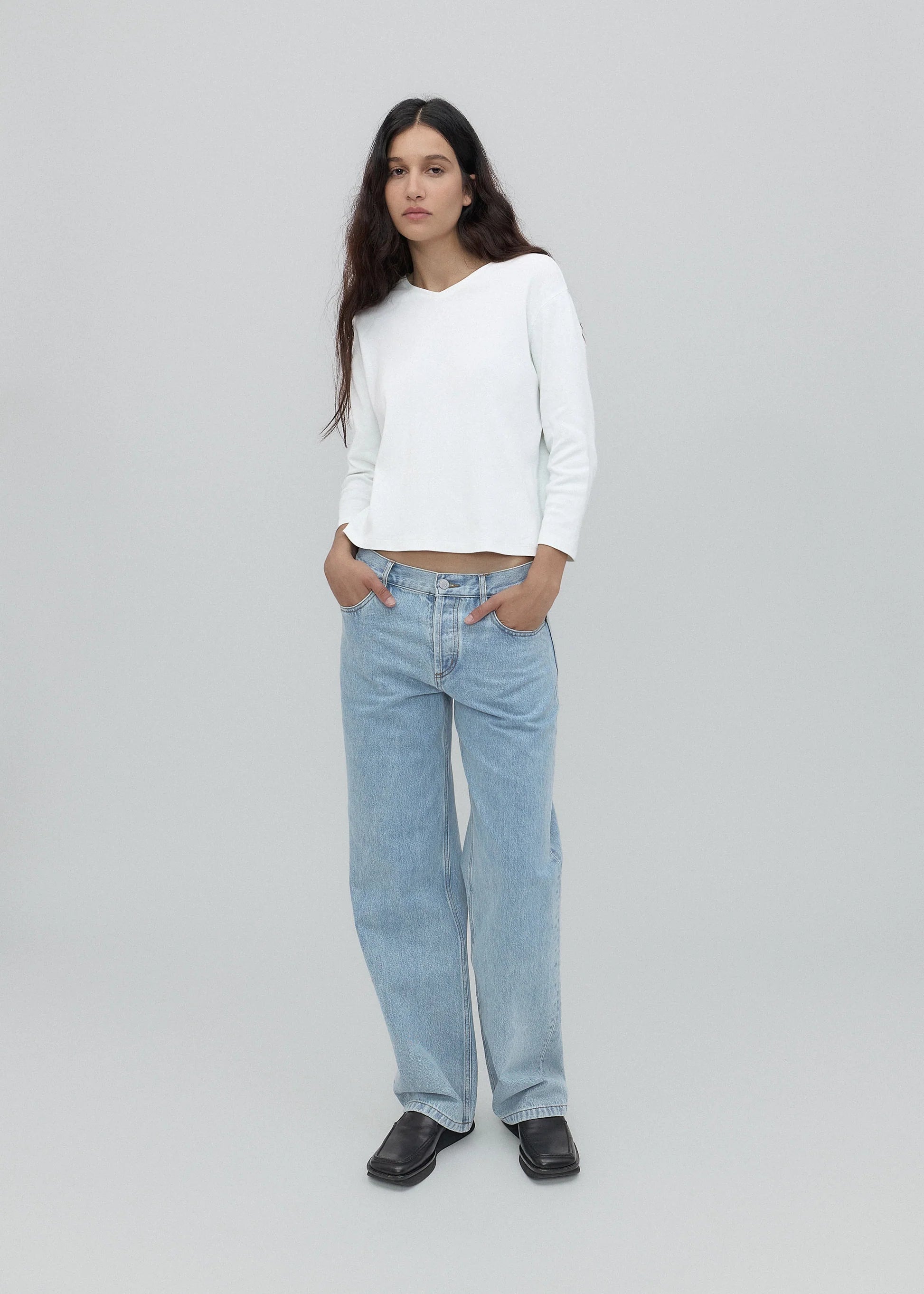 A person with long dark hair stands against a plain light gray background, wearing a white long-sleeve shirt, black shoes, and the Everyday Jeans in light blue—a loose fit perfect for everyday wear—with hands in their pockets.