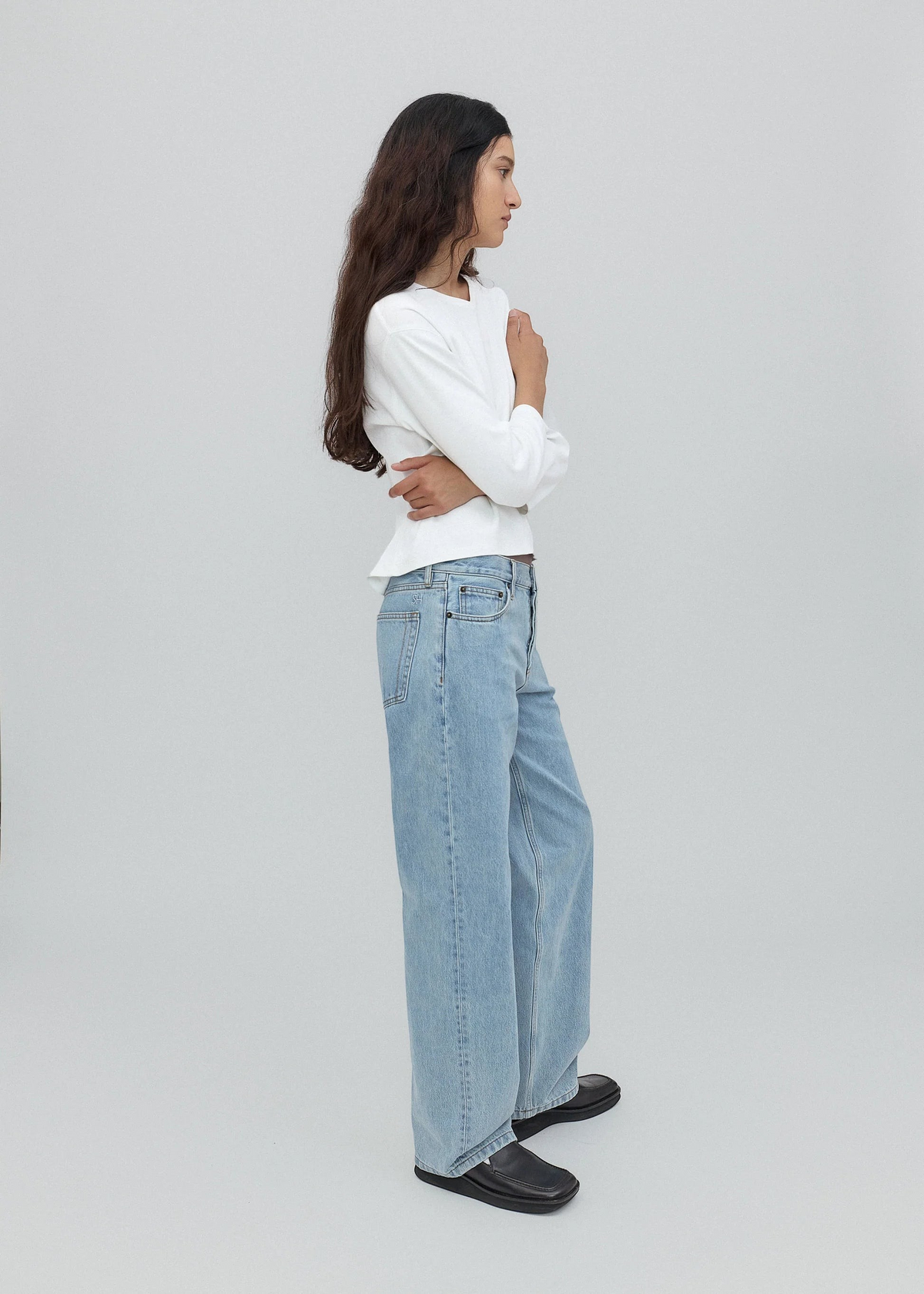 A woman with long dark hair stands in profile, wearing a white long-sleeve shirt, light blue Everyday Jeans featuring a relaxed silhouette, and black shoes against a plain light gray background.