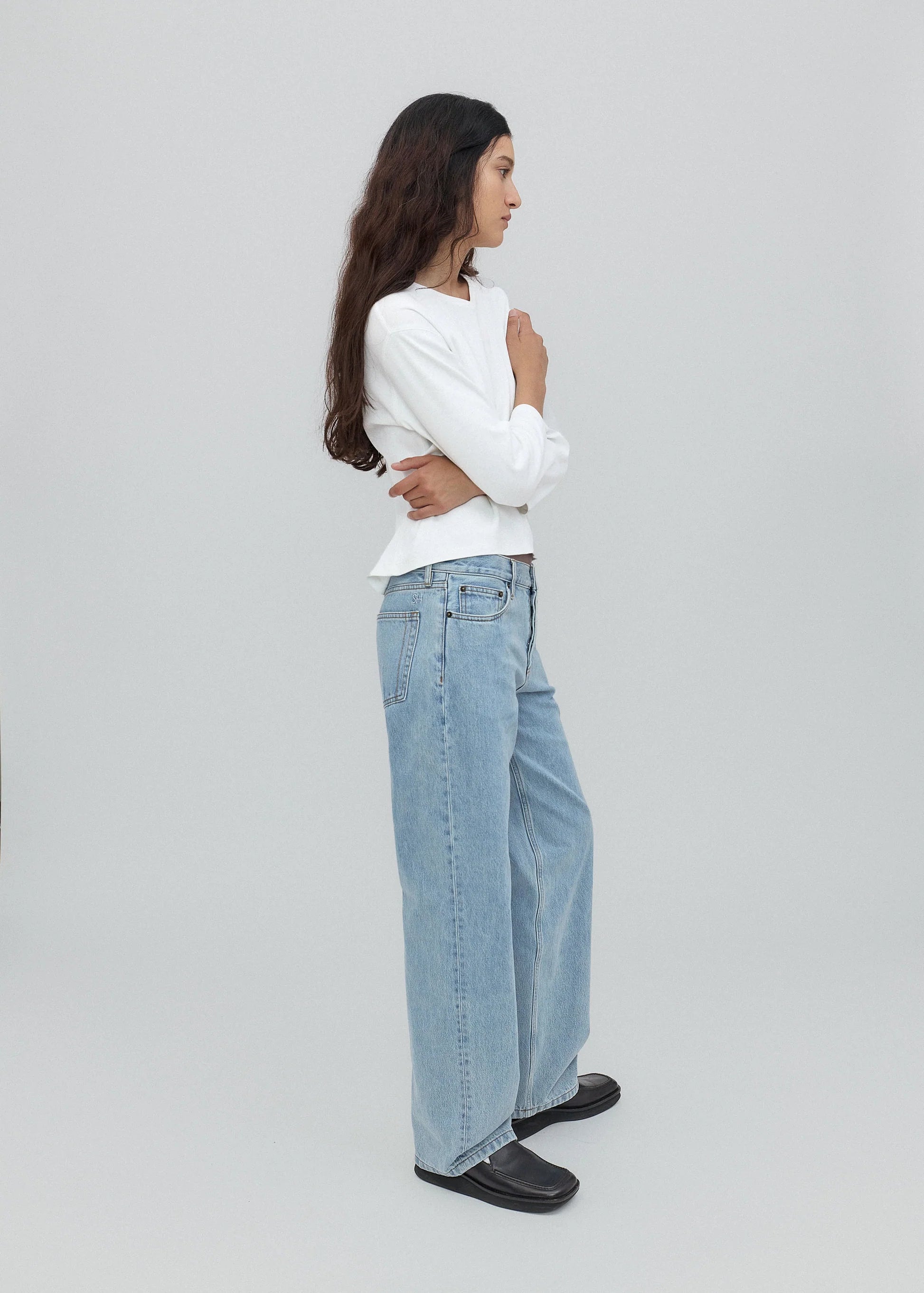 A woman with long dark hair stands in profile, wearing a white long-sleeve shirt, light blue Everyday Jeans featuring a relaxed silhouette, and black shoes against a plain light gray background.
