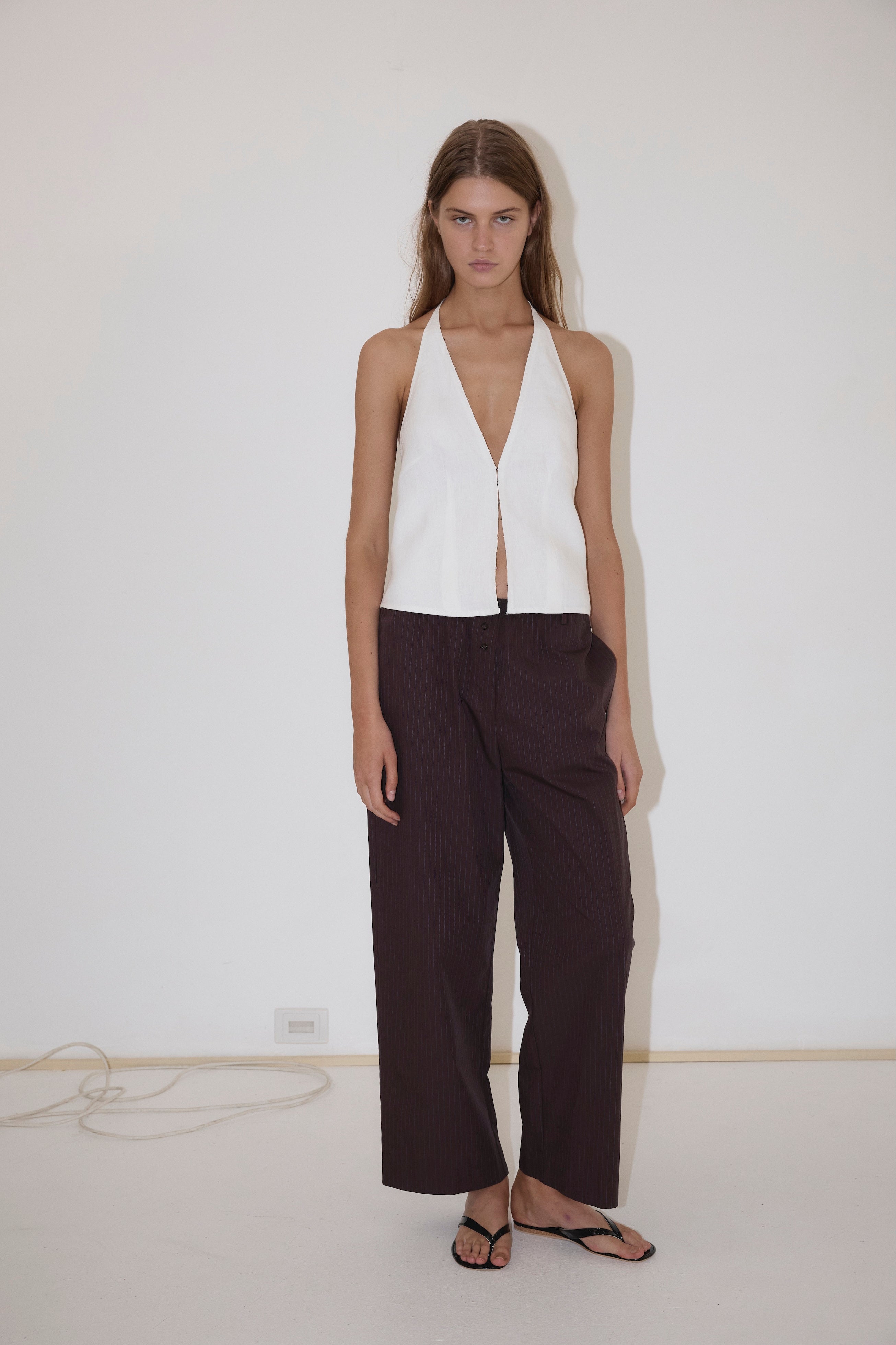 A woman stands against a white wall wearing the Close Halter Top from Deiji Studios, paired with loose dark brown pinstripe pants and black flip-flops. A cable is visible on the floor along the wall.
