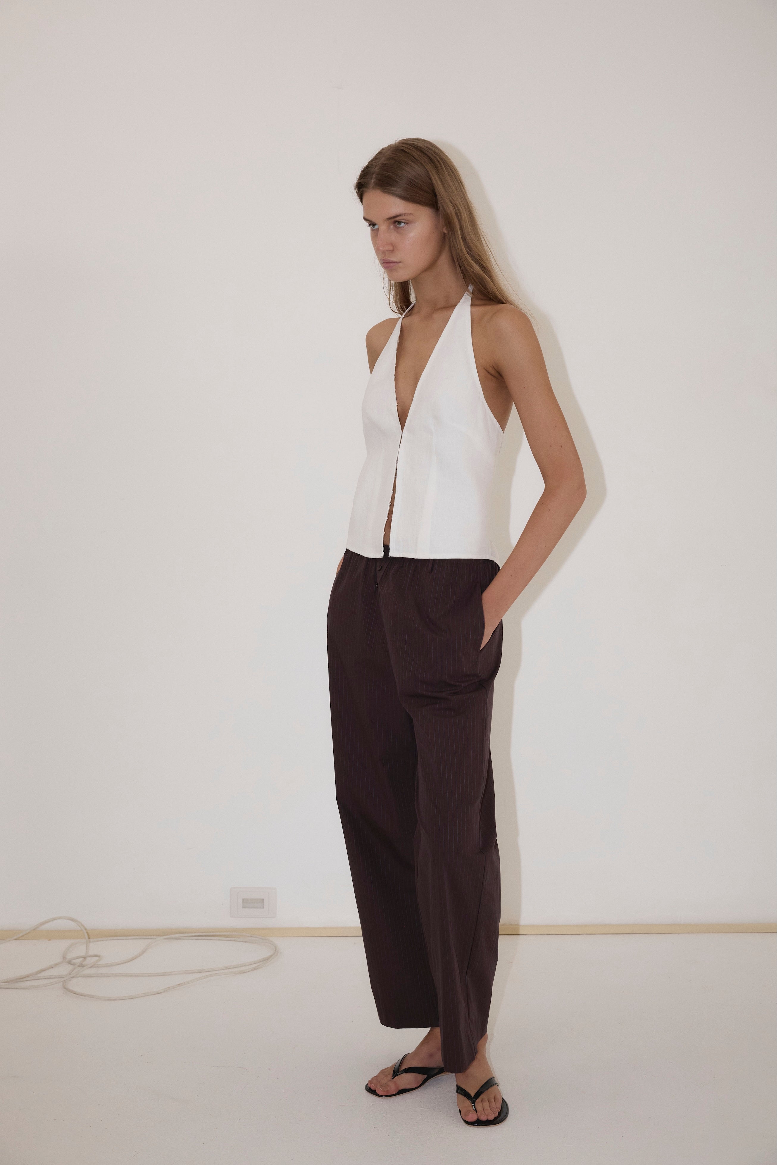 A person stands against a white wall, wearing the Close Halter Top from Deiji Studios—a sleeveless white halter top—paired with loose dark brown pants and black sandals, with hands in their pockets.