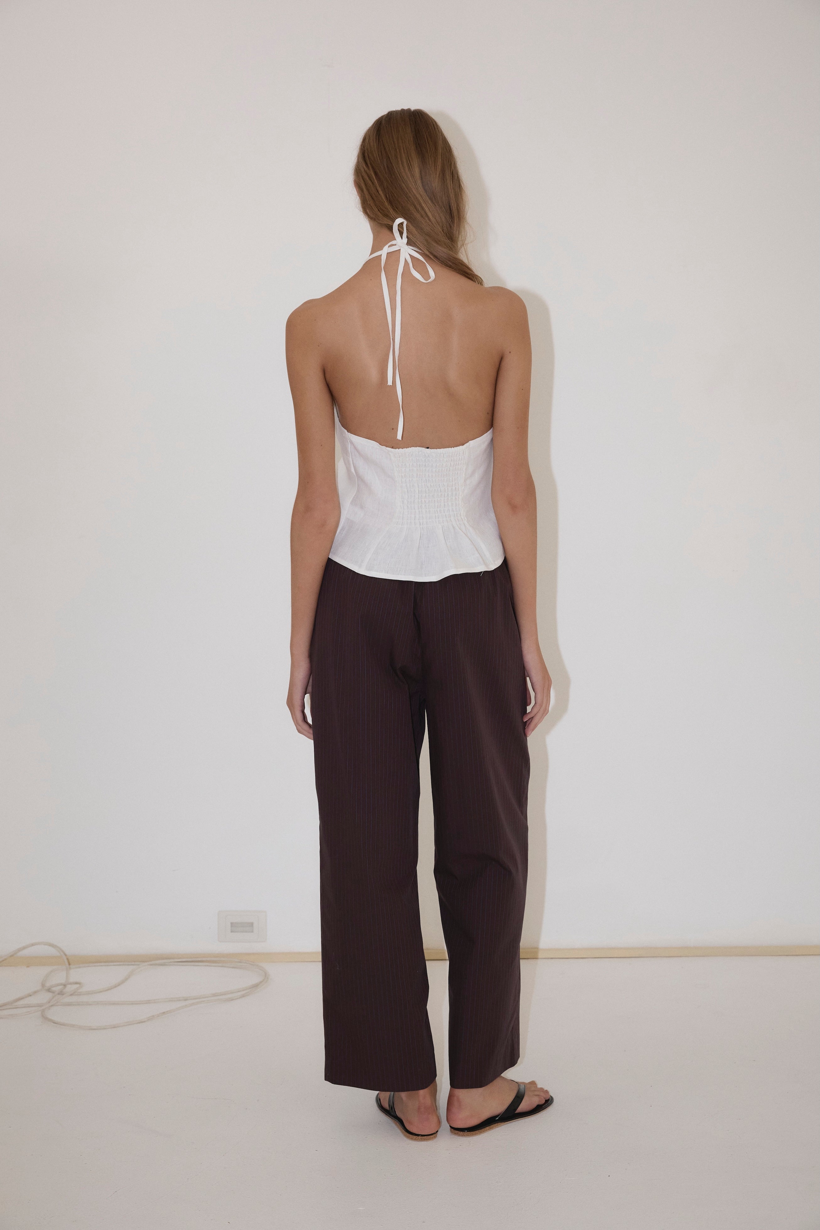 A person stands facing a white wall, wearing the Close Halter Top from Deiji Studios, paired with loose dark pants and black sandals. Their hair is down, and a power cord is visible on the floor near the wall.