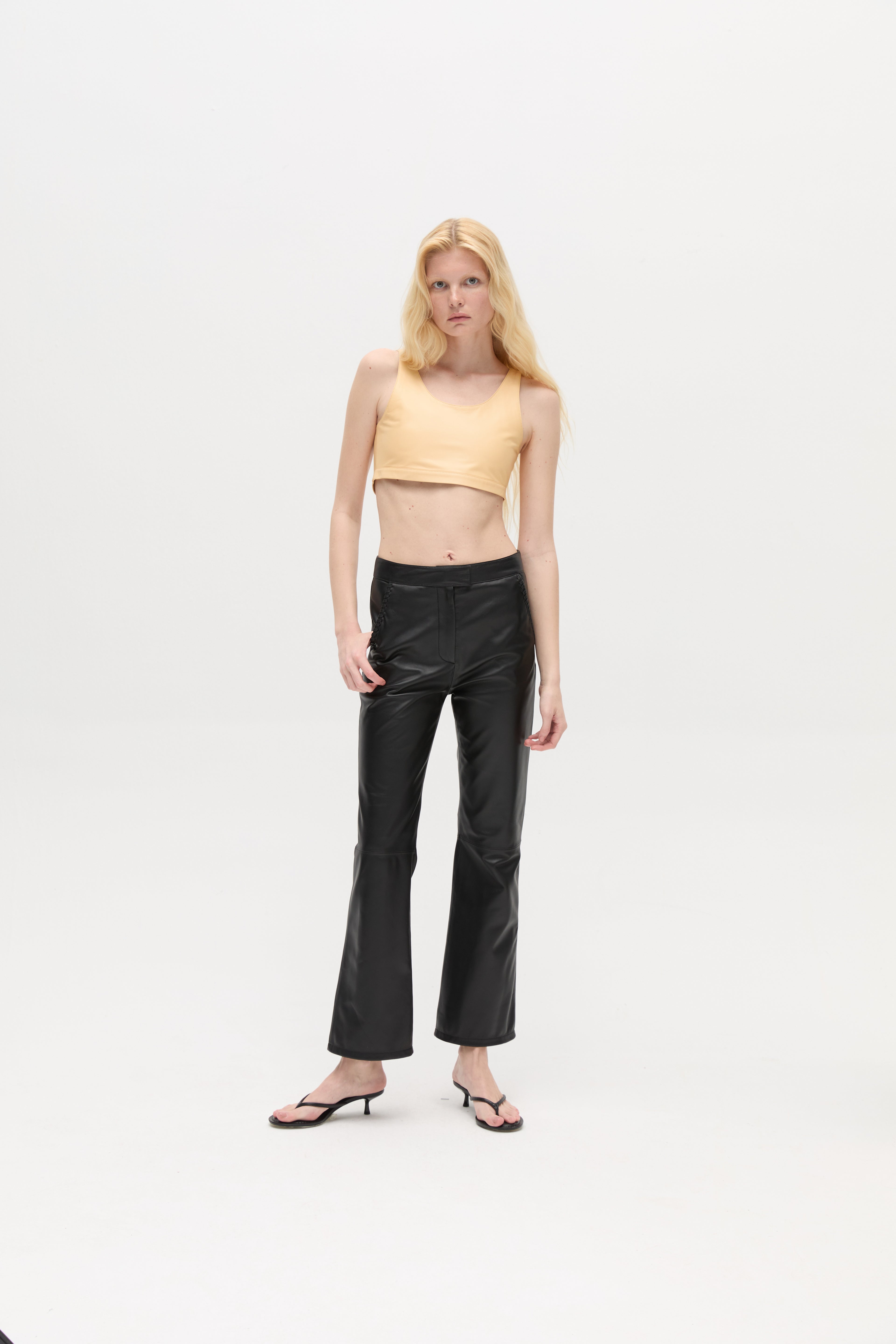 A woman with long blonde hair wears a beige crop top, the Xena Pants in black leather, and black sandals, standing against a plain white background.