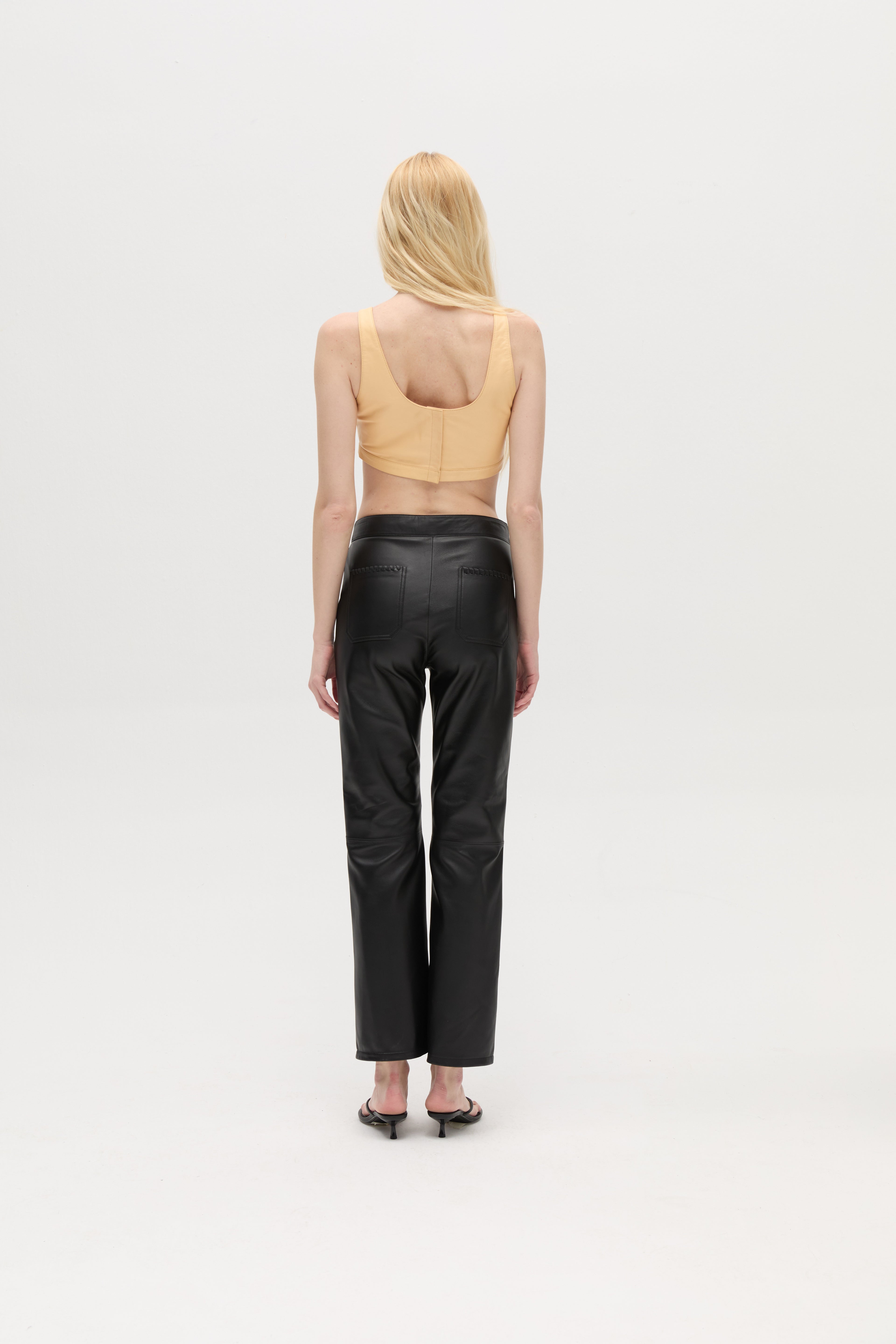 A person with long blonde hair is standing facing away, wearing a peach sleeveless top, the Xena Pants in black leather, and black heeled sandals against a plain white background.