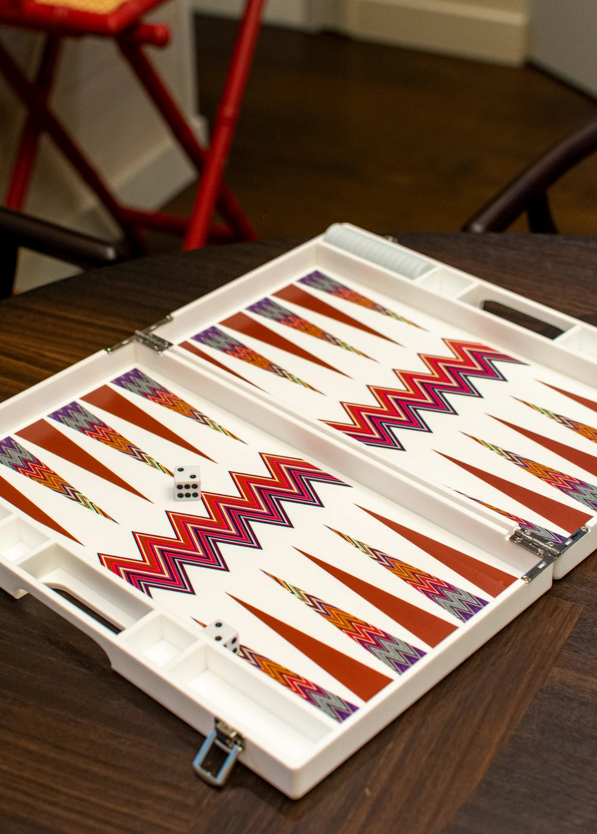 An open ZigZag Backgammon Board Game with a colorful design sits on a wooden coffee table. The set’s vibrant acrylic accents catch the light, while two dice rest on the board and part of a red chair is visible in the background.
