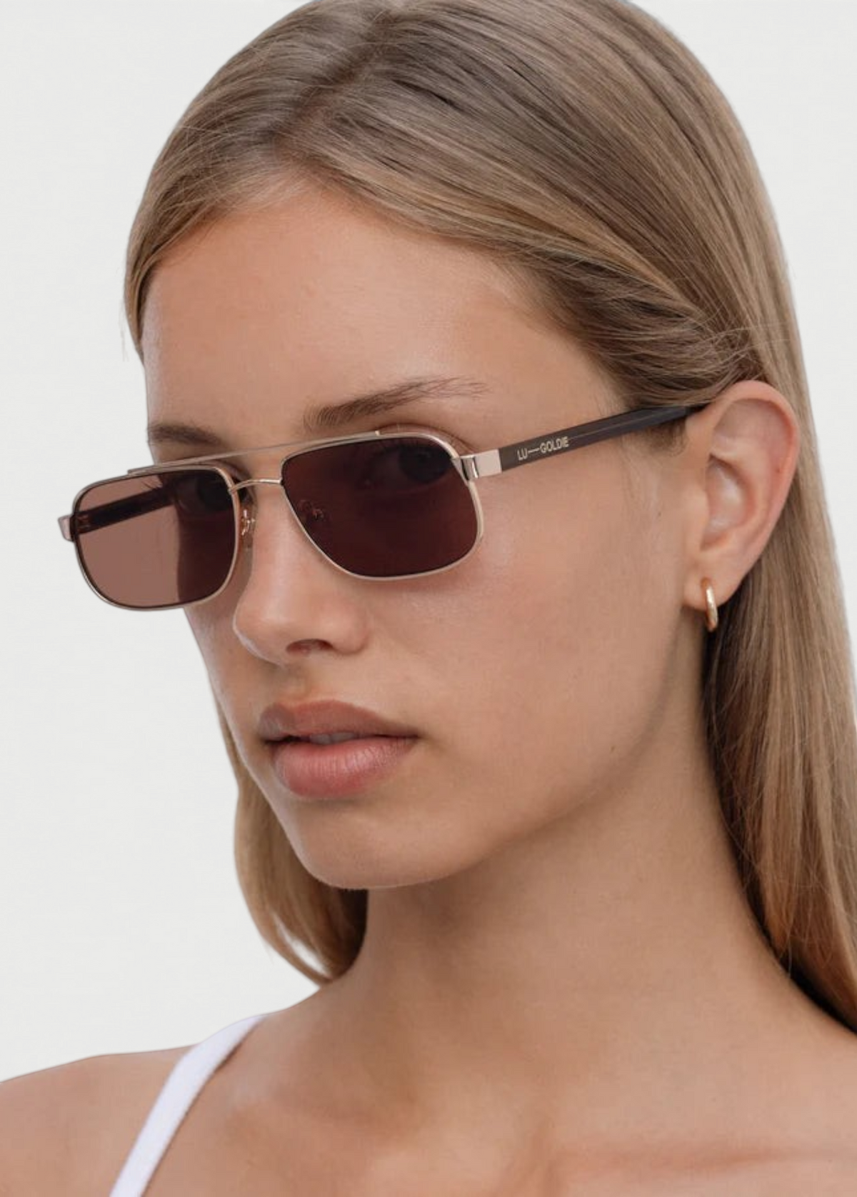 A woman with straight, light brown hair wears the Evangeline rectangular brown sunglasses, which feature a stainless steel frame and is styled with a small gold hoop earring. She has a neutral expression and is dressed in a white top against a plain background.