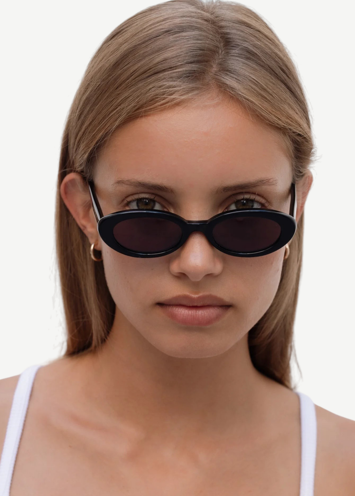 A young woman with straight light brown hair wears the Sylvie sunglasses in classic black and a white tank top, looking directly at the camera against a plain white background.