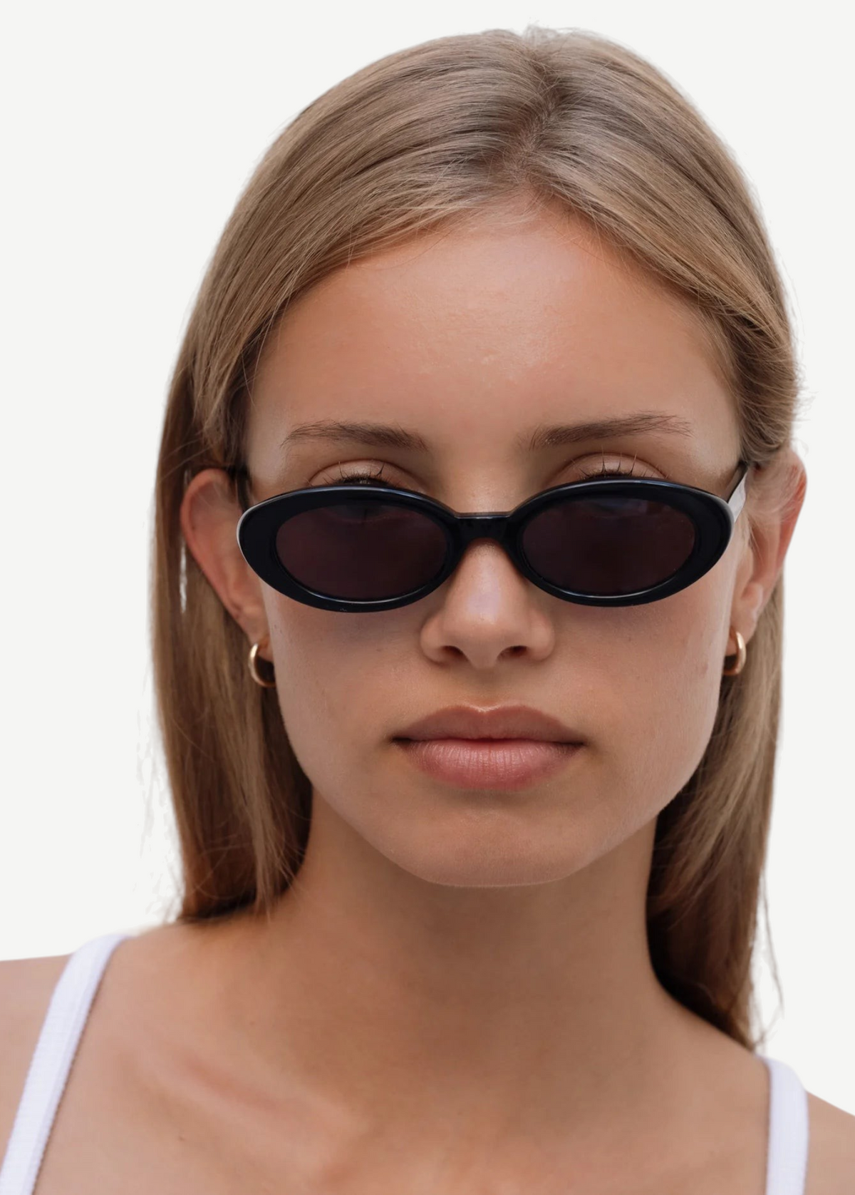 A person with straight, light brown hair wears the Sylvie sunglasses in black, paired with small hoop earrings and a white top, looking directly at the camera against a plain light background.