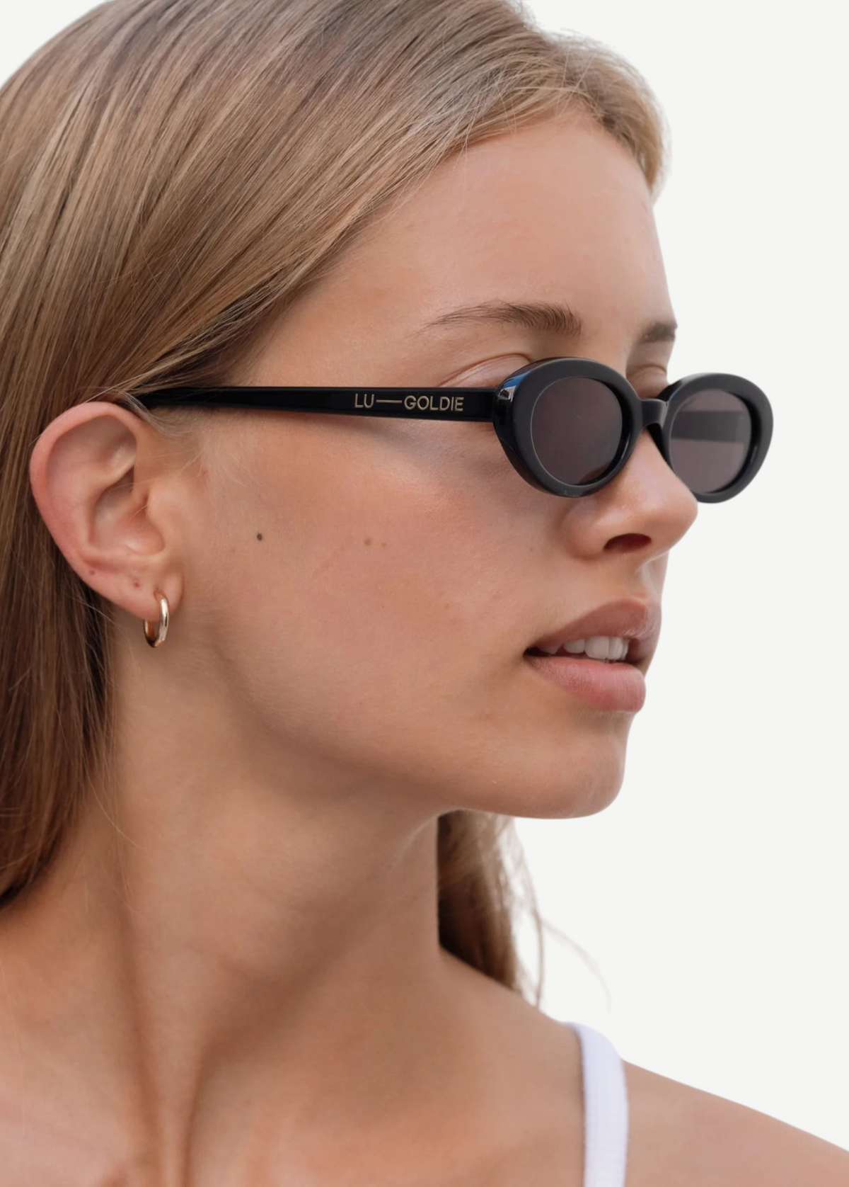 A woman with straight, light brown hair wears the Sylvie sunglasses in black, paired with a gold hoop earring and a white tank top—her effortlessly chic look enhanced as she glances to the side against a plain, light background.