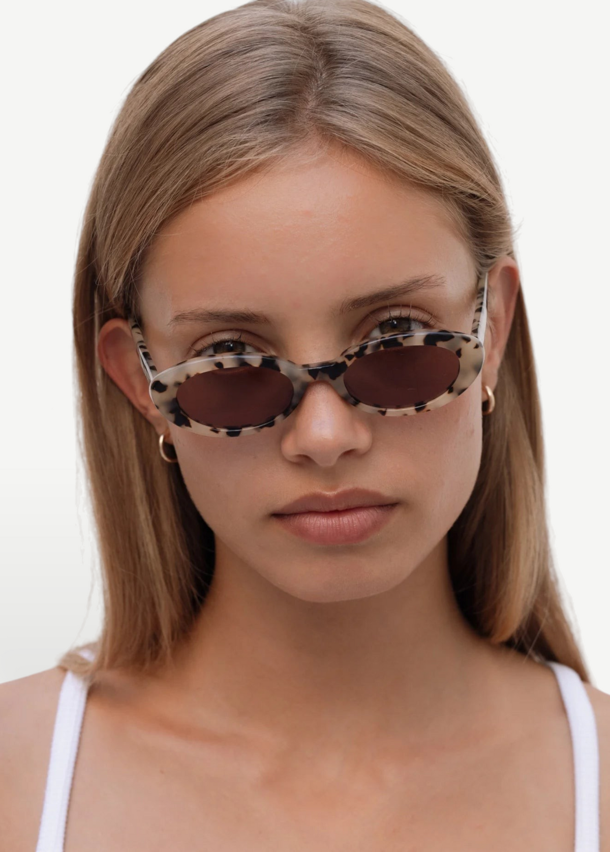 A woman with long blonde hair wears the Sylvie sunglasses—featuring an oval shape, a spotted frame, and dark UV400 lenses—along with small hoop earrings. She pairs them with a white tank top while posing against a plain white background.