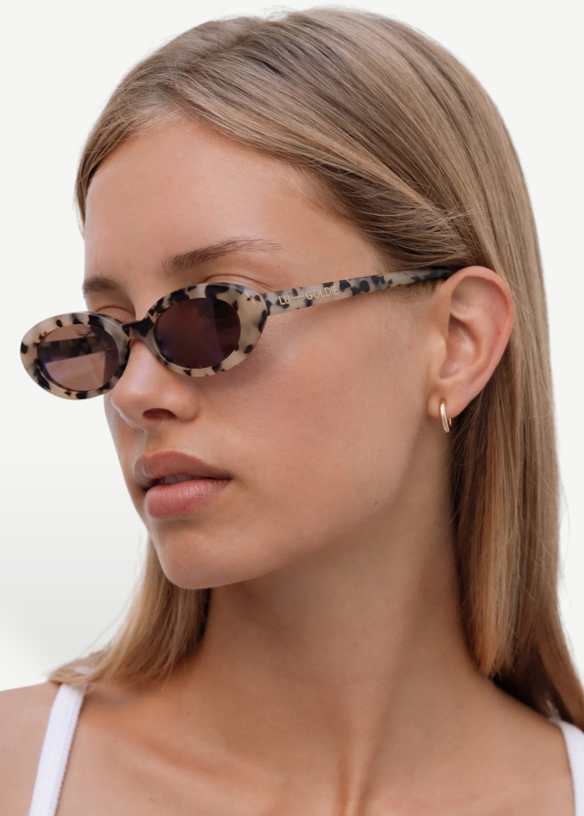 A woman with straight, light brown hair wears Sylvie oval tortoiseshell sunglasses with UV400 protection and a small gold hoop earring. She has a neutral expression and is dressed in a white top against a plain white background.
