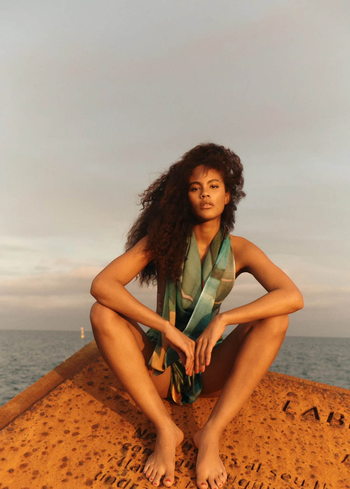 KABE SHOP INC Magdalena Pareo with a fluid landscape print worn by a woman sitting on a rusted structure by the sea.