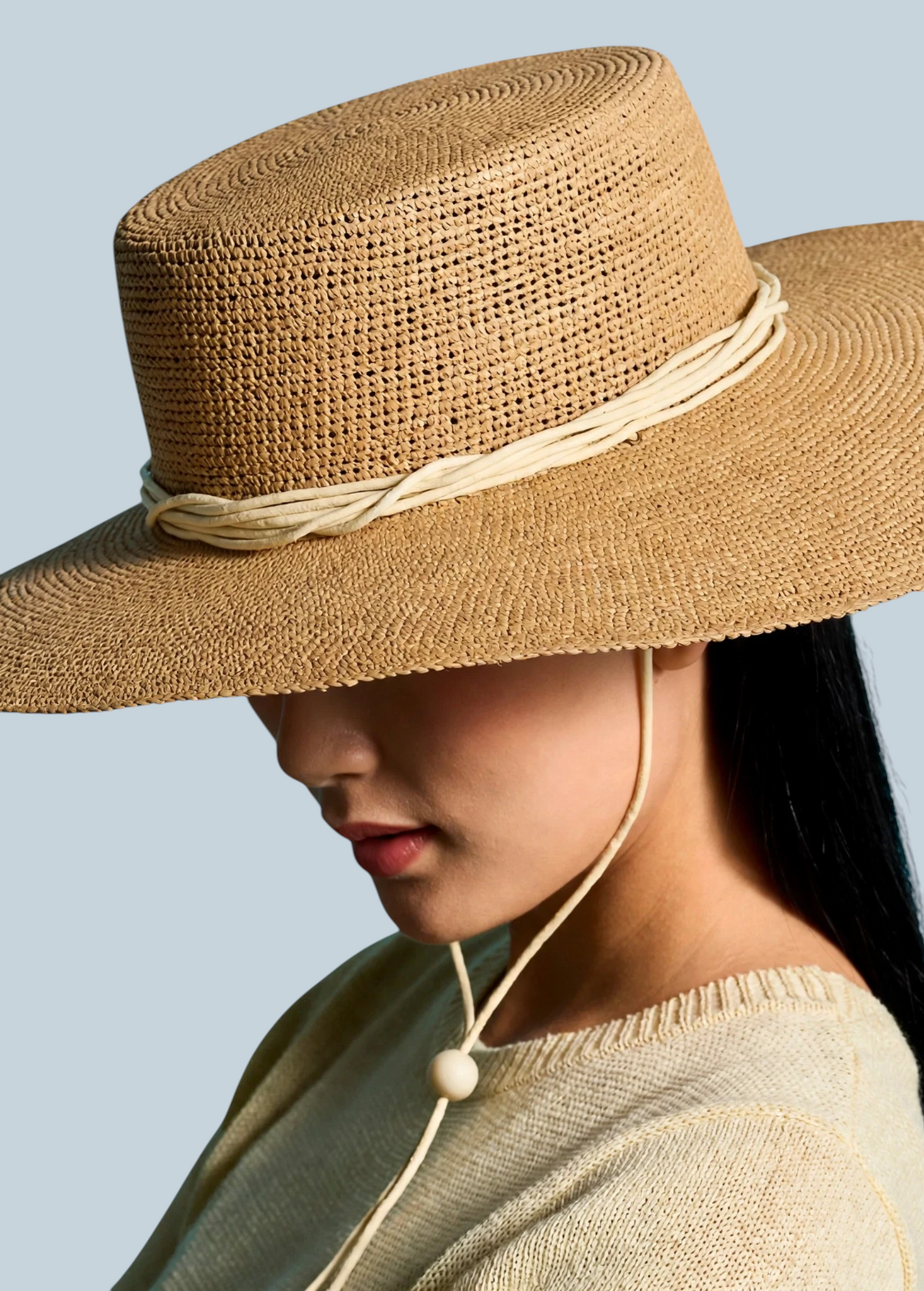 KABE SHOP INC Vitoria wide brim straw hat in tan with a cream cord chin strap worn by a woman against a blue backdrop