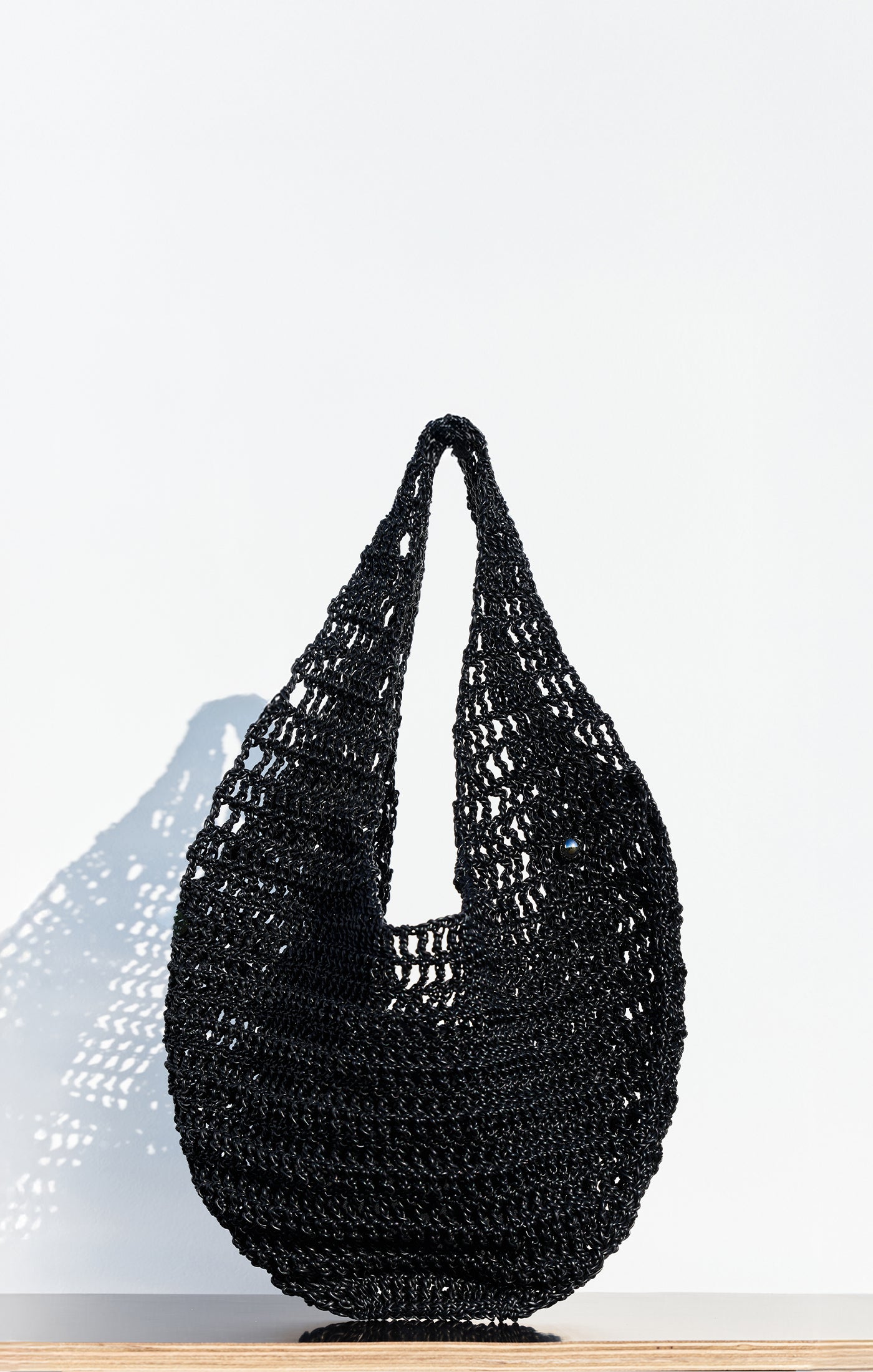 The Market Net Bag, a black, woven crochet-style handbag with a single looped handle, sits on a light wooden surface against a plain white background.