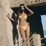 A woman in a yellow eco-friendly swimwear set stands by a textured stone structure, touching her hair. Sunlight highlights her and a palm frond is visible in the foreground. The background shows blue sky and water, while the adjustable side tie Praia Bottom, crafted from Peach Perfect fabric, adds both comfort and style to her look.