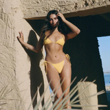 A woman in a yellow eco-friendly swimwear set stands by a textured stone structure, touching her hair. Sunlight highlights her and a palm frond is visible in the foreground. The background shows blue sky and water, while the adjustable side tie Praia Bottom, crafted from Peach Perfect fabric, adds both comfort and style to her look.
