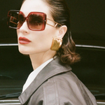 A woman with slicked-back hair wears the oversized square Macy Sunglasses, which feature gold plated temples, along with bold gold earrings, red lipstick, and a dark leather jacket as she stands in front of a brown vintage car.
