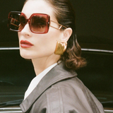 A woman with slicked-back hair wears the oversized square Macy Sunglasses, which feature gold plated temples, along with bold gold earrings, red lipstick, and a dark leather jacket as she stands in front of a brown vintage car.