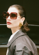 A woman with slicked-back hair wears the oversized square Macy Sunglasses, which feature gold plated temples, along with bold gold earrings, red lipstick, and a dark leather jacket as she stands in front of a brown vintage car.