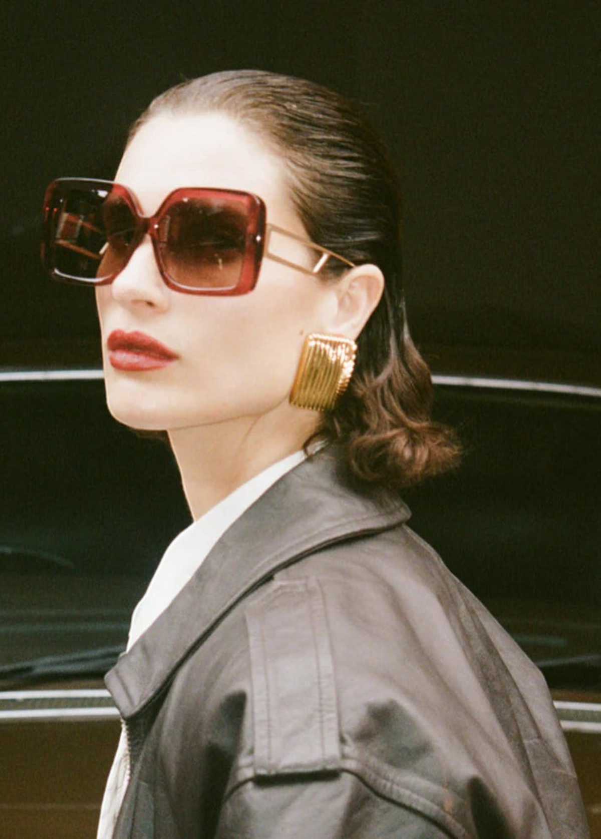 A woman with slicked-back hair wears the oversized square Macy Sunglasses, which feature gold plated temples, along with bold gold earrings, red lipstick, and a dark leather jacket as she stands in front of a brown vintage car.