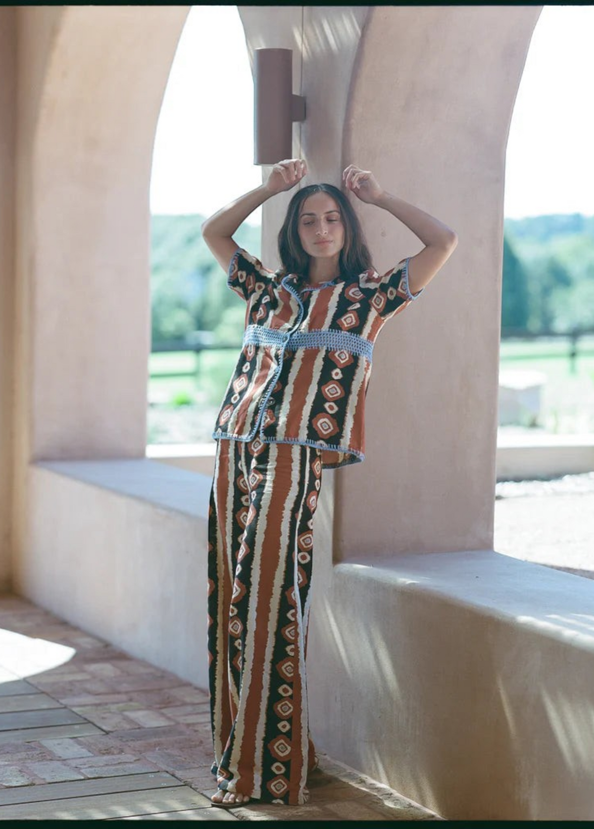 Woman wearing KABE SHOP INC Nera wide leg pants with a matching ikat print shirt in a sunlit arched corridor.