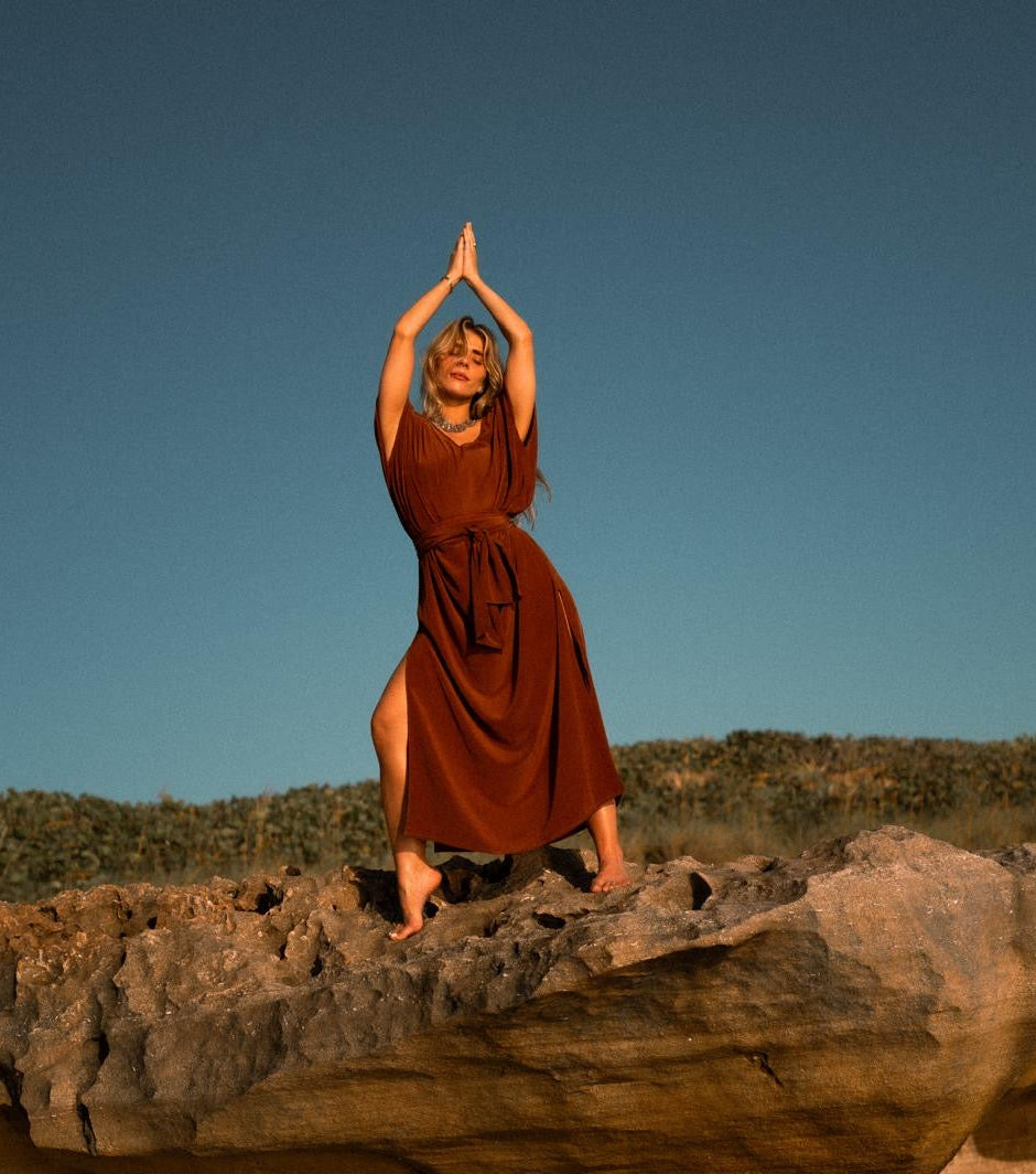 A person in a rust-colored, elegant silk kaftan stands barefoot on a rock, arms raised and palms together above their head, their relaxed silhouette framed by shrub-covered hills and a clear blue sky in the background.