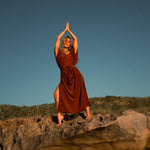 A person in a rust-colored, elegant silk kaftan stands barefoot on a rock, arms raised and palms together above their head, their relaxed silhouette framed by shrub-covered hills and a clear blue sky in the background.
