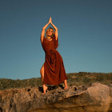 A person in a rust-colored, elegant silk kaftan stands barefoot on a rock, arms raised and palms together above their head, their relaxed silhouette framed by shrub-covered hills and a clear blue sky in the background.