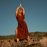A person in a rust-colored, elegant silk kaftan stands barefoot on a rock, arms raised and palms together above their head, their relaxed silhouette framed by shrub-covered hills and a clear blue sky in the background.