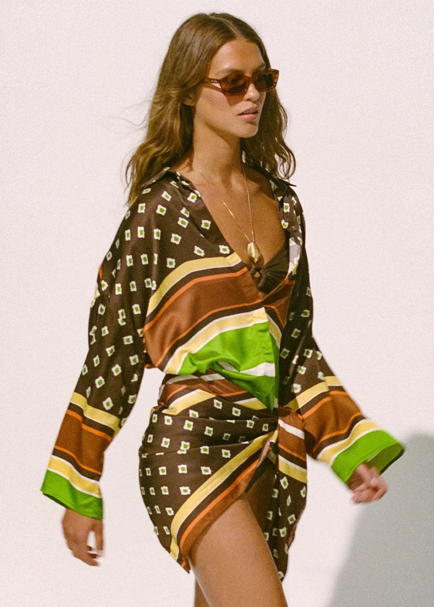 A person wearing a long-sleeved, geometric-patterned Cora Shirt, knotted at the waist over a bikini top.