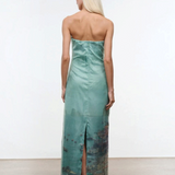 Viewed from behind, a person wears the Amara Dress, a strapless, mint-green maxi featuring a photographic print of a coastal city at the hem and a back slit.