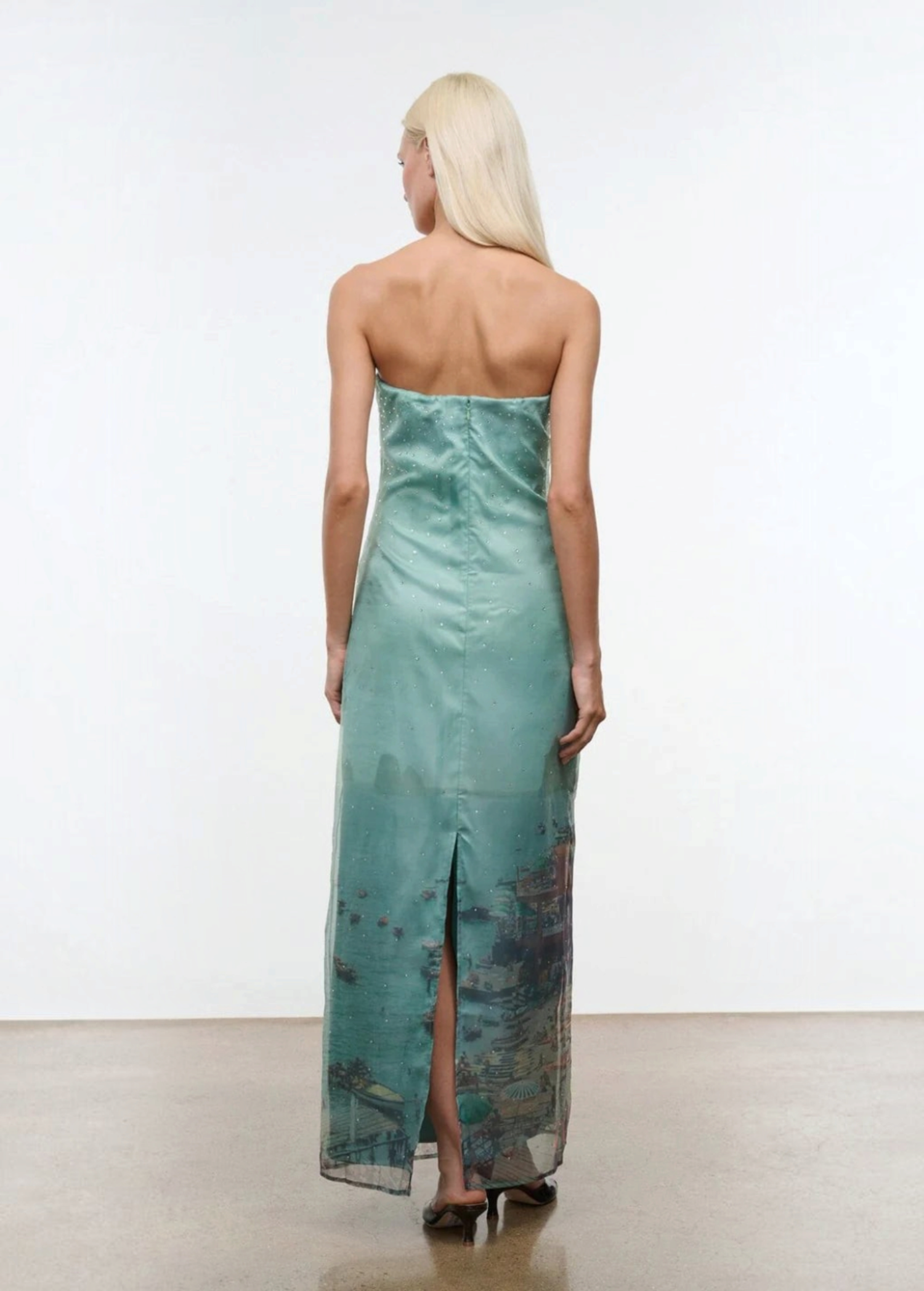 Viewed from behind, a person wears the Amara Dress, a strapless, mint-green maxi featuring a photographic print of a coastal city at the hem and a back slit.