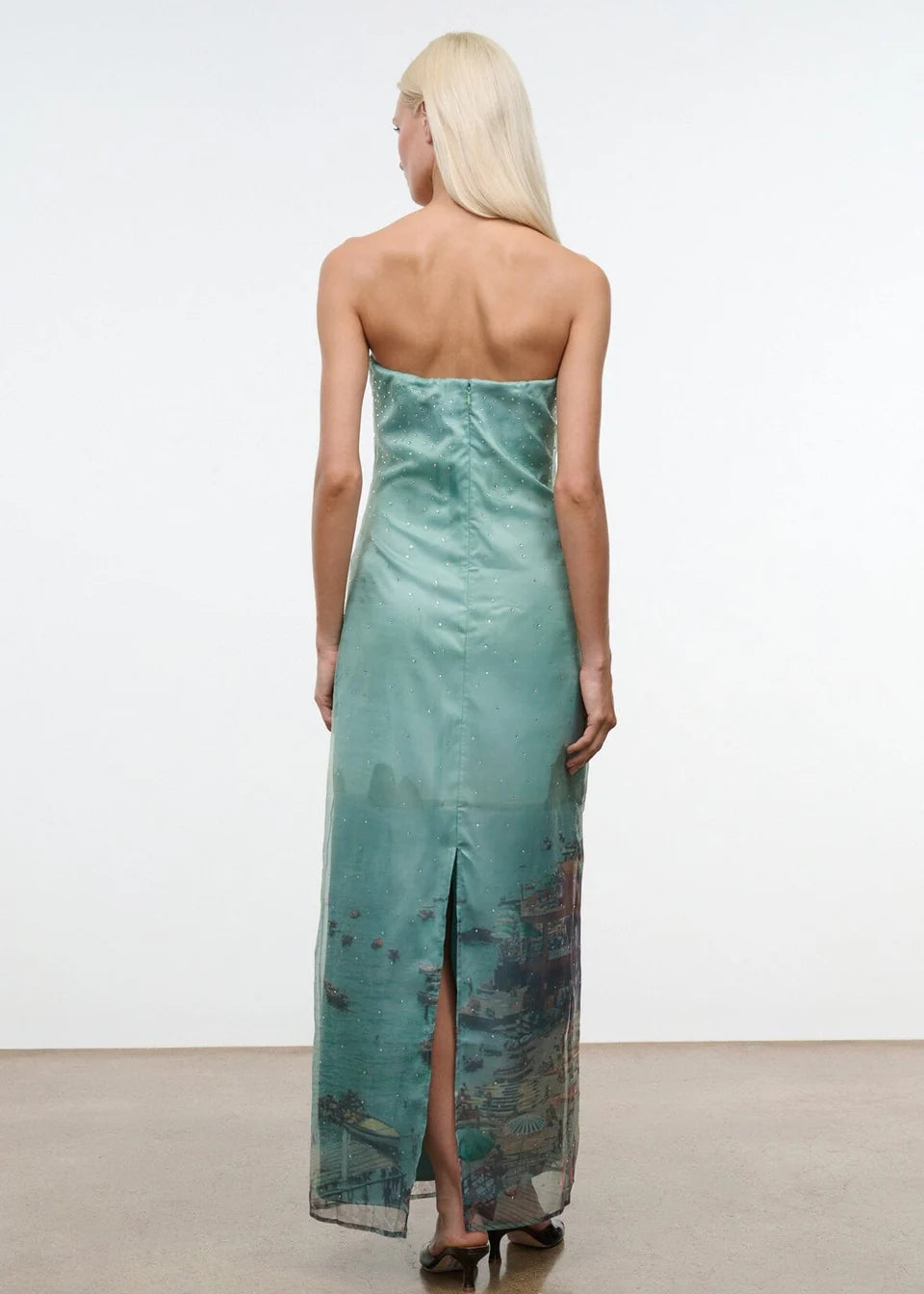 Viewed from behind, a person wears the Amara Dress, a strapless, mint-green maxi featuring a photographic print of a coastal city at the hem and a back slit.