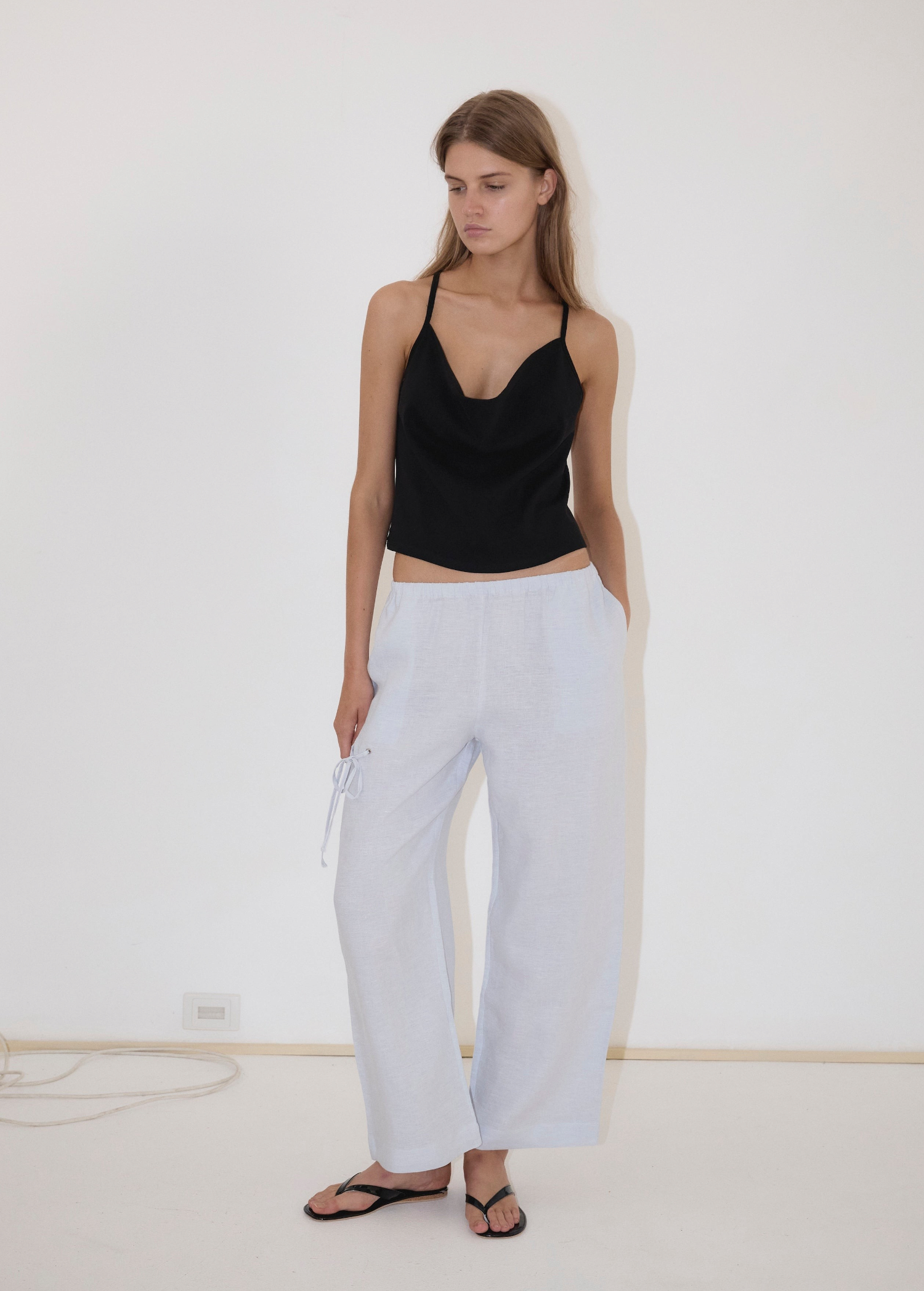 A person wearing a black camisole and light blue Deiji Studios Eyelet Pant, featuring a side drawstring and a straight-leg silhouette.
