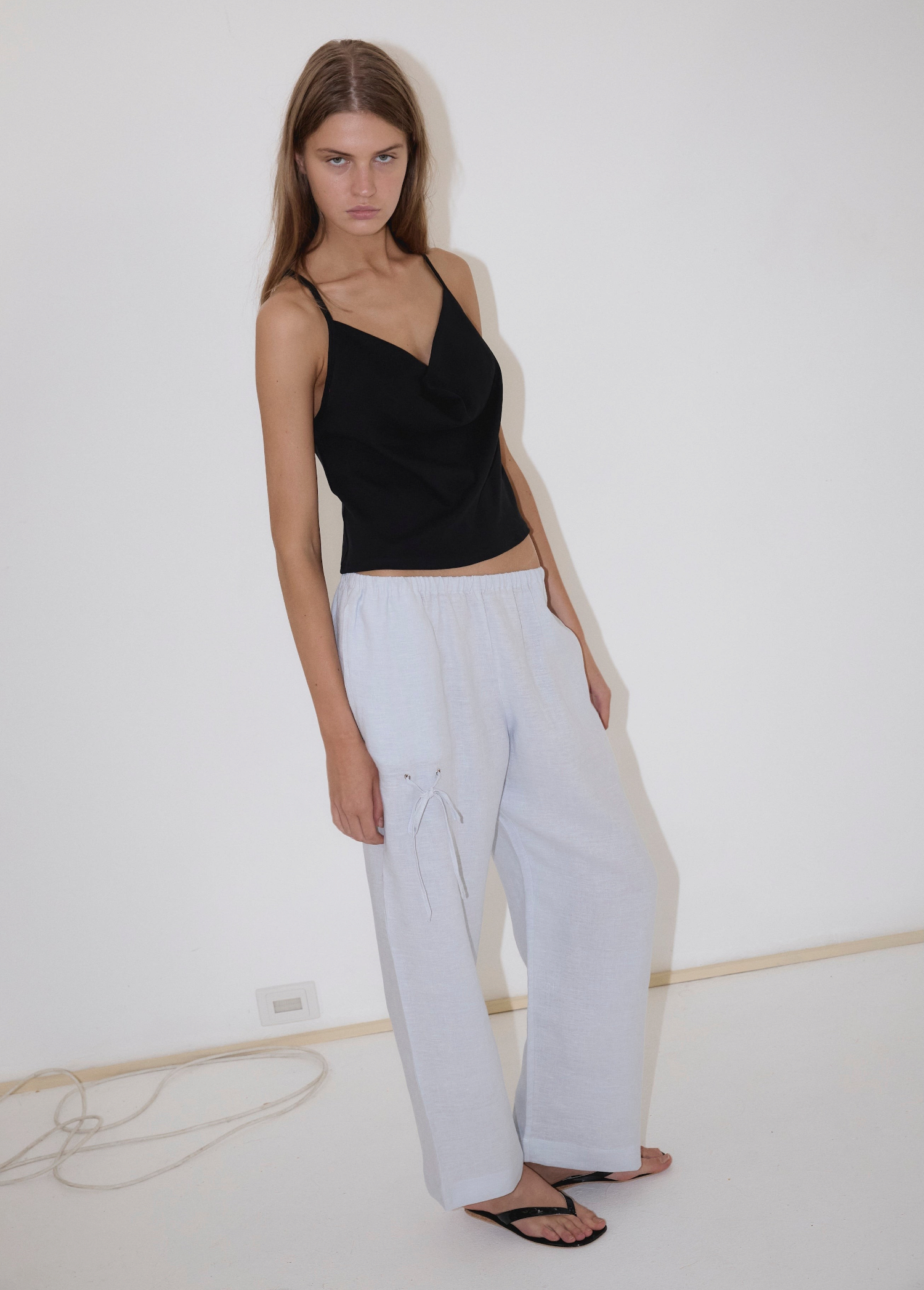 A person wearing a black camisole and white Eyelet Pant in a straight-leg silhouette, featuring loose-fitting fabric and a tied drawstring detail at the hip.
