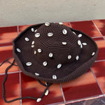 The Wika Hat Shells, a handmade brown crochet hat crafted from organic cotton and adorned with white cowrie shells, rests on a red-brown tiled surface against a beige wall.