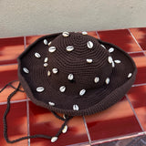 The Wika Hat Shells, a handmade brown crochet hat crafted from organic cotton and adorned with white cowrie shells, rests on a red-brown tiled surface against a beige wall.