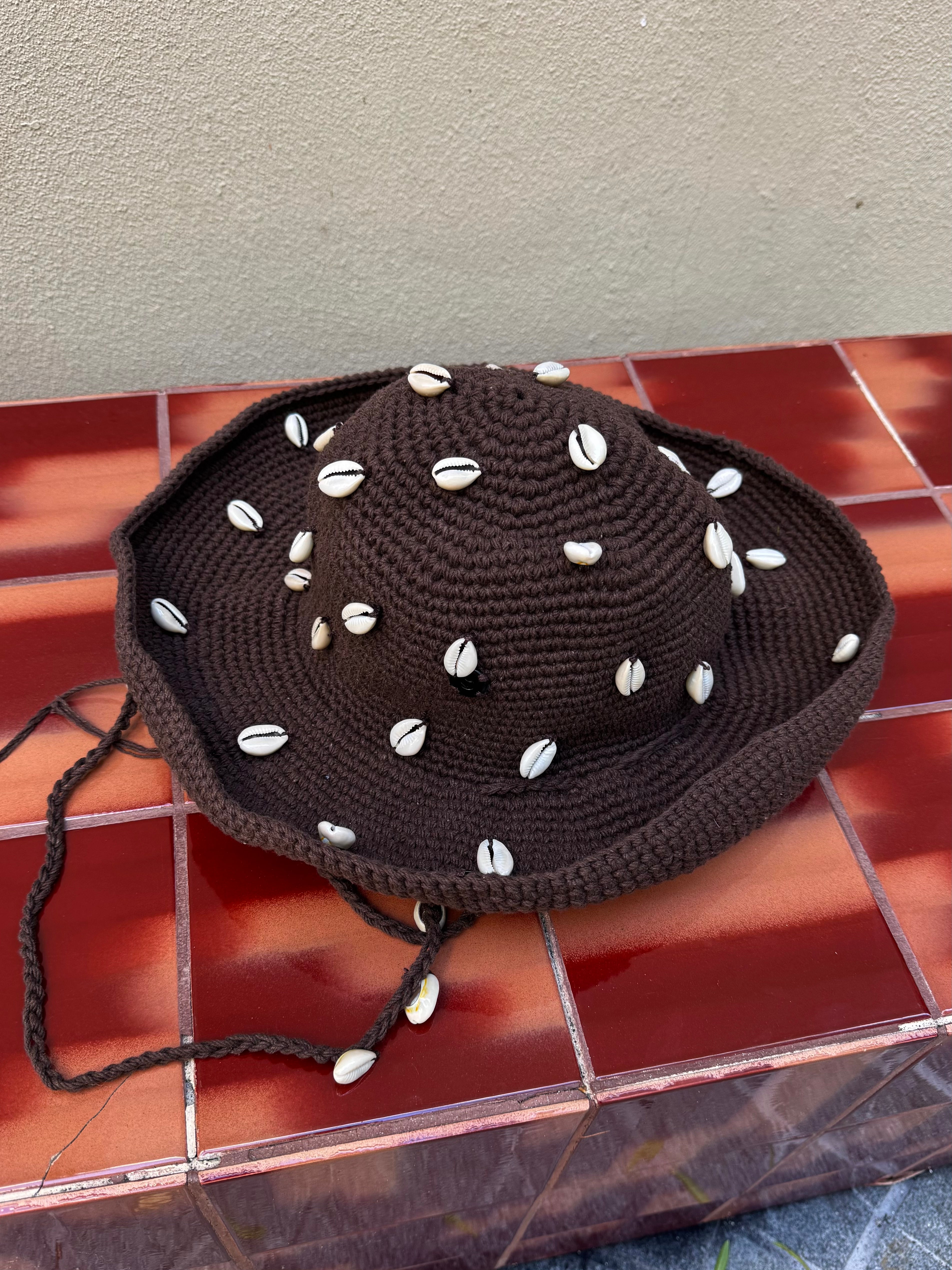The Wika Hat Shells, a handmade brown crochet hat crafted from organic cotton and adorned with white cowrie shells, rests on a red-brown tiled surface against a beige wall.
