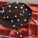 The Wika Hat Shells, a handmade brown crochet hat crafted from organic cotton and adorned with white cowrie shells, rests on a red-brown tiled surface against a beige wall.