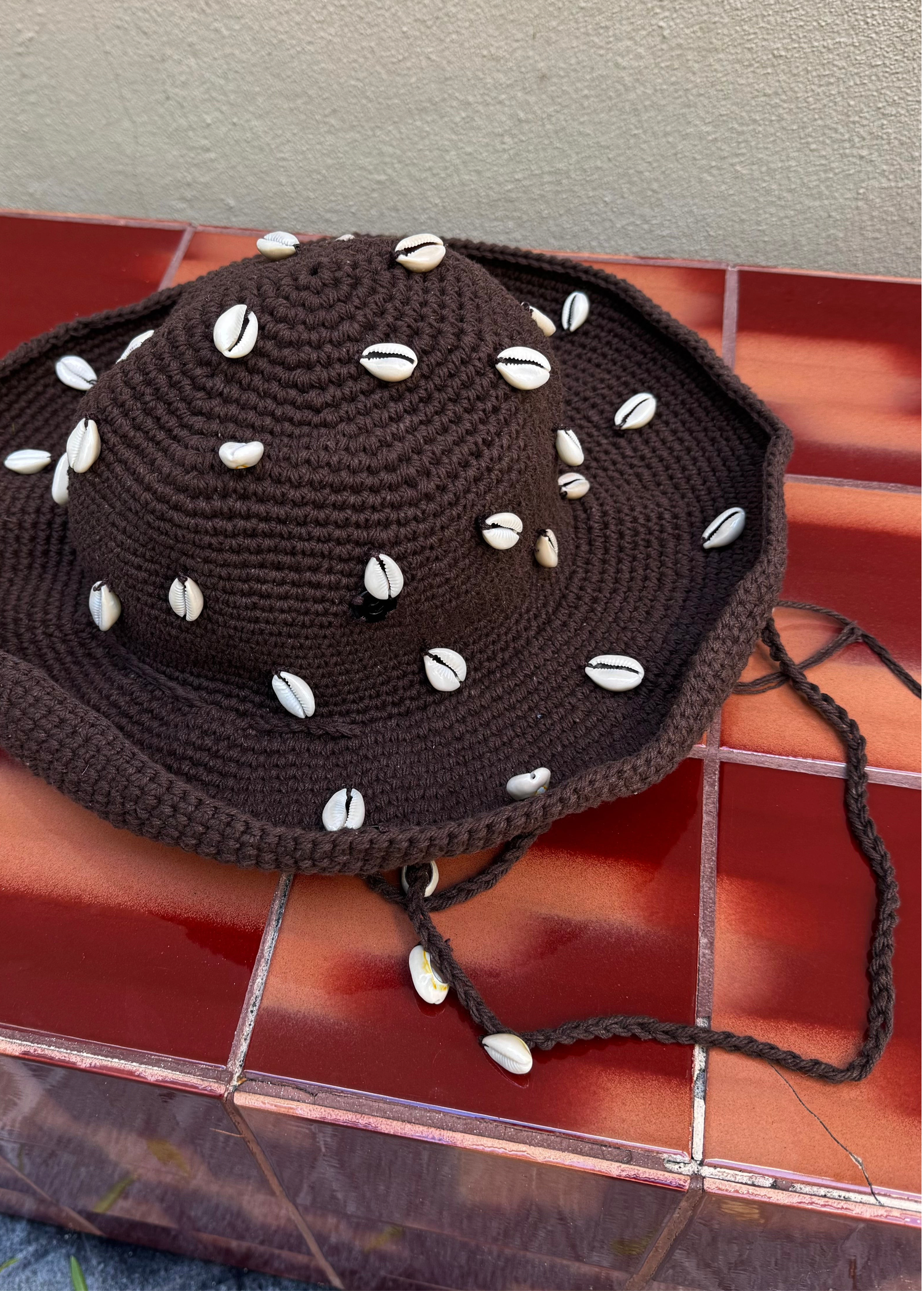 The Wika Hat Shells, a handmade brown crochet hat crafted from organic cotton and adorned with white cowrie shells, rests on a red-brown tiled surface against a beige wall.
