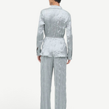 A person seen from behind wears a matching long-sleeved button-up shirt and wide-leg Nami Pant in a shimmering, crinkled sage green fabric, creating a relaxed silhouette.