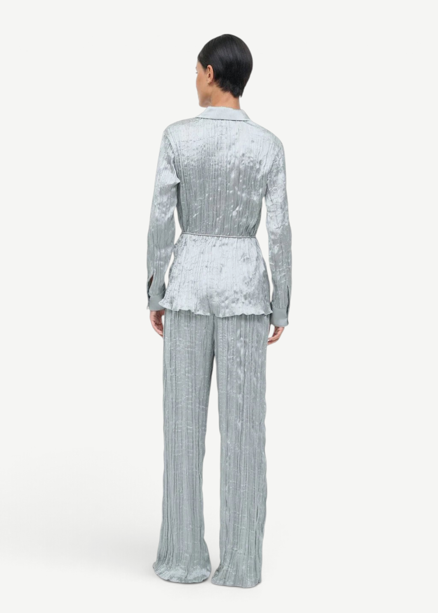 A person seen from behind wears a matching long-sleeved button-up shirt and wide-leg Nami Pant in a shimmering, crinkled sage green fabric, creating a relaxed silhouette.