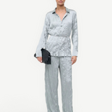 A model wears the Suki Shirt, a textured light gray long-sleeved button-down with a matching thin waist tie and wide-leg trousers, styled effortlessly with a black woven clutch.