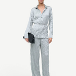 A person wears a crinkled light blue long-sleeve button-down shirt and matching Nami Pant in a relaxed silhouette, holding a black woven clutch.