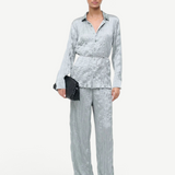 A person wears a crinkled light blue long-sleeve button-down shirt and matching Nami Pant in a relaxed silhouette, holding a black woven clutch.