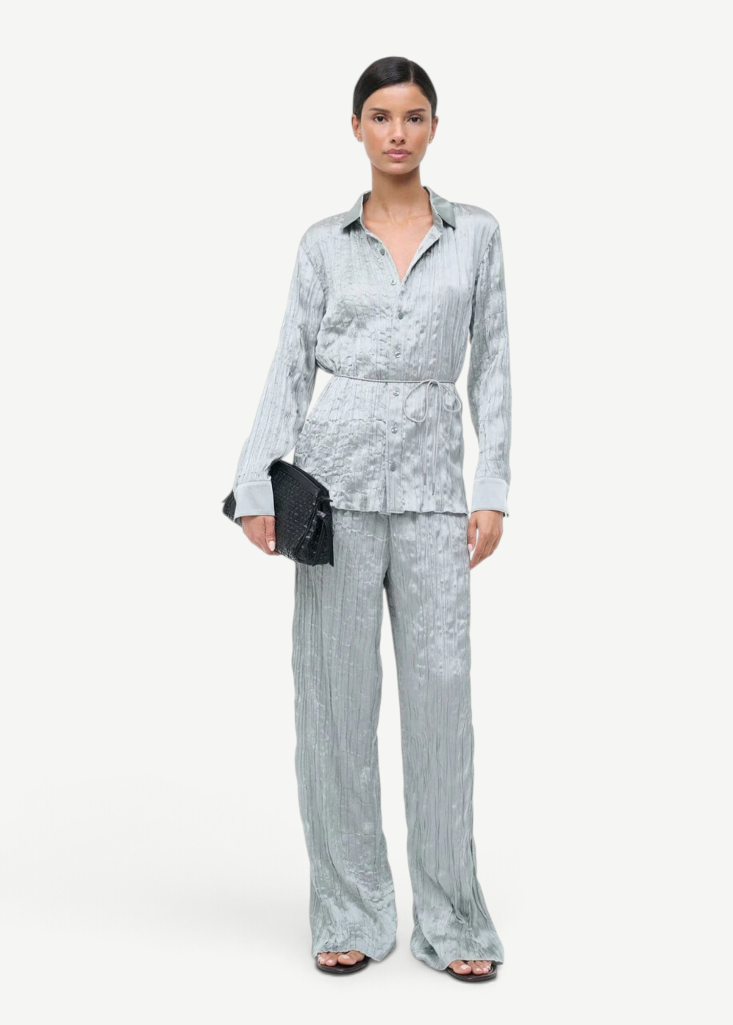 A person wears a crinkled light blue long-sleeve button-down shirt and matching Nami Pant in a relaxed silhouette, holding a black woven clutch.