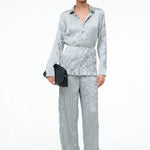 A person wears a crinkled light blue long-sleeve button-down shirt and matching Nami Pant in a relaxed silhouette, holding a black woven clutch.