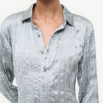A person wearing the Suki Shirt, a sage green button-up with a long-sleeved, crinkled texture that serves as a versatile wardrobe essential.